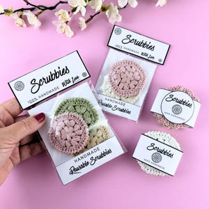 May include: Handmade reusable scrubbies in various colors, including pink, green, and cream, are displayed on a pink background. Each scrubbie is packaged with a label that reads "Scrubbies 100% Handmade With Love."