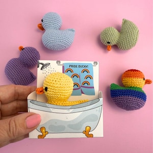 Peut inclure: Plusieurs canards crochetés faits à la main de différentes couleurs, dont bleu, violet, vert, jaune et arc-en-ciel. Un canard est dans une carte en forme de baignoire avec le texte "Pride Ducky!" et des serviettes arc-en-ciel. Les canards ont des becs oranges sur fond rose.