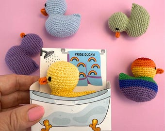 Puede incluir: Una colección de patos de ganchillo hechos a mano en varios colores, incluyendo azul, morado, verde, amarillo y arcoíris. Un pato está en una tarjeta con forma de bañera con el texto "Pride Ducky!" y toallas de arcoíris. Los patos tienen picos naranjas sobre un fondo rosa.