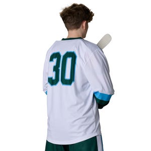 May include: A white hockey jersey featuring the number 30 in dark green with a light blue outline on the back. The long-sleeved jersey has dark green and light blue bands on the sleeves and is paired with dark green shorts. A hockey stick is visible.