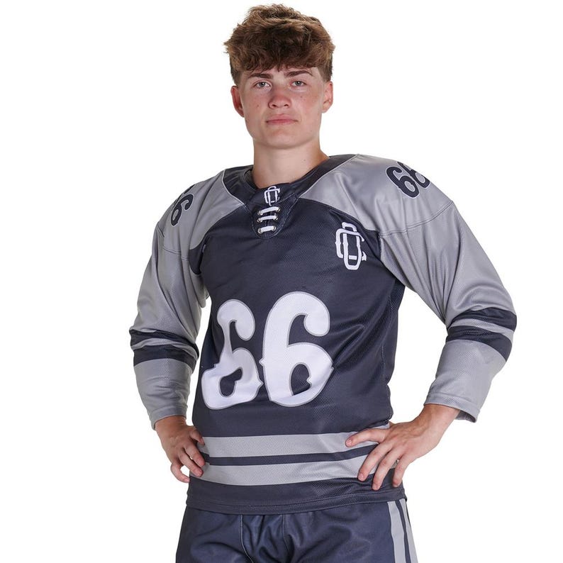 Puede incluir: Una camiseta de hockey de color azul marino oscuro y gris con el n&uacute;mero 66 en fuente blanca grande en el pecho y los hombros. La camiseta incluye un detalle de cuello con cordones, un logotipo blanco en el pecho y dos franjas grises horizontales.