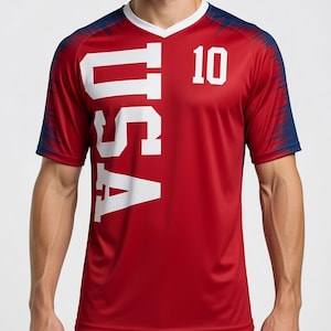 May include: Red athletic jersey with the word "USA" in large white letters and the number "10". The jersey has a white V-neck collar and navy blue accents on the shoulders and sleeves. The jersey is made of a breathable, athletic material.