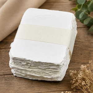 May include: A stack of rectangular white paper sheets with rough, deckled edges. The stack is secured in the centre by a wide, smooth, light beige paper band that wraps horizontally around the middle of the white paper collection.