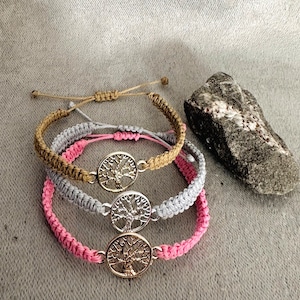 Può includere: Tre braccialetti intrecciati con chiusure regolabili, ognuno con un ciondolo ad albero della vita argentato. I braccialetti sono in tonalità marrone, grigio e rosa.
