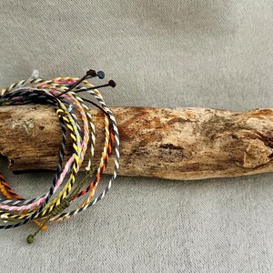 Könnte beinhalten: Eine Sammlung bunter geflochtener Armbänder mit schwarzen und weißen Streifen, rosa und weißen Streifen, gelben und weißen Streifen sowie grünen und weißen Streifen. Die Armbänder sind zusammengebunden und auf einem Stück Treibholz ausgestellt.