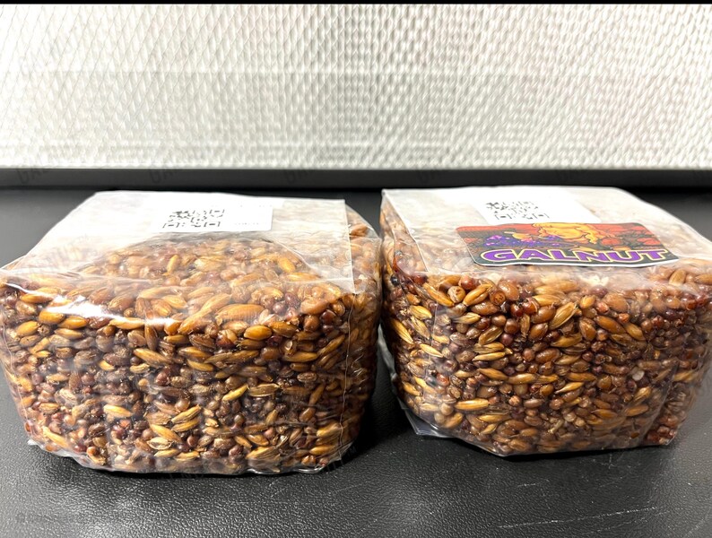 May include: Two clear plastic bags filled with brown, oval-shaped grains. Each bag has a white label on top, and the right bag features a rectangular sticker with the text GALNUT in purple letters and a graphic of a yellow, round character.