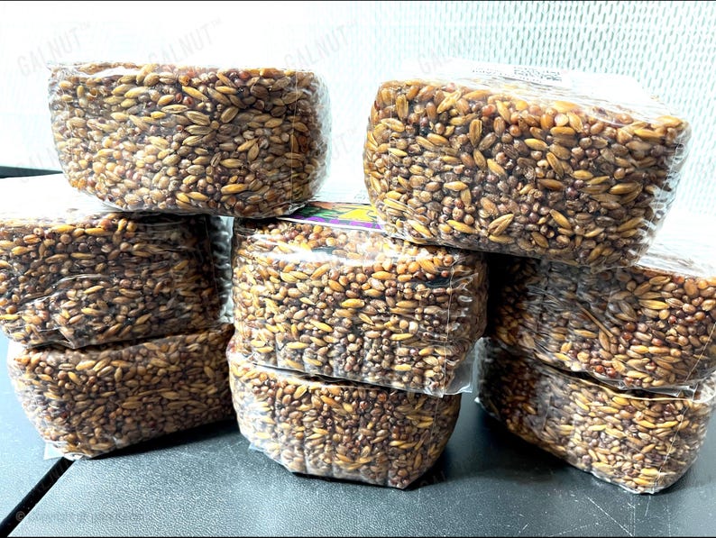 May include: A collection of seven rectangular blocks of compressed brown grains, each individually wrapped in clear plastic film. The grains vary in shades of tan, golden brown, and dark brown, creating a textured, dense appearance.