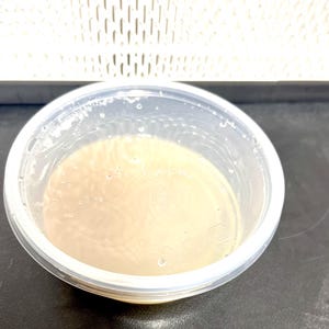May include: A clear, round plastic container filled with a light brown liquid. The container has a slightly opaque appearance and is sitting on a dark surface. The liquid has a smooth texture with small bubbles.