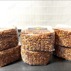 May include: Five rectangular blocks of compressed brown grains, individually wrapped in clear plastic. Each block is densely packed with a mixture of light and dark brown seeds and grains. A small rectangular label with a graphic is visible on top.