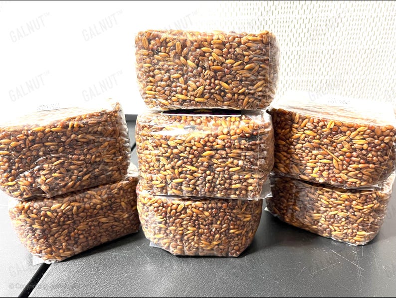 May include: Five rectangular, clear plastic-wrapped packages containing a dense mixture of brown and golden-brown grains. The packages are stacked in a pyramid formation, with the text GALNUT printed repeatedly on the transparent packaging.