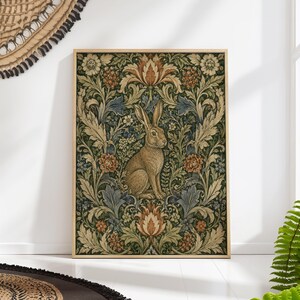 Könnte beinhalten: Ein gerahmter Kunstdruck mit einem braunen Hasen vor einem dunklen floralen Muster. Das Kunstwerk zeigt ein detailliertes Design mit Grün-, Blau- und Orangetönen, das eine Vintage-Ästhetik erzeugt. Der Rahmen ist hellholzfarben.