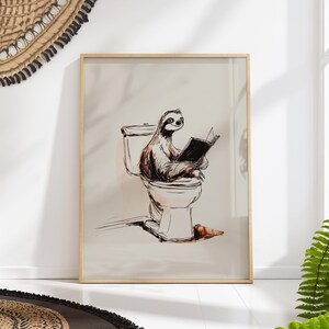 Peut inclure: Une affiche encadrée présente une illustration fantaisiste d'un paresseux lisant sur des toilettes. L'œuvre d'art est dans des tons bruns et blancs, avec un fond beige clair et un cadre en bois.