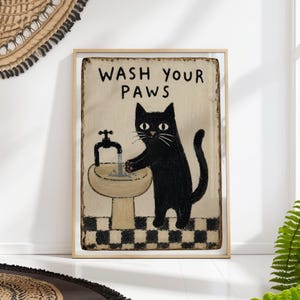 Peut inclure: Tableau encadré représentant un chat noir se lavant les pattes à un évier. Le texte "WASH YOUR PAWS" est au-dessus du chat. L'évier est beige et le sol est un motif en damier noir et blanc. L'œuvre est dans un cadre en bois.