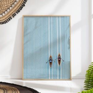 Puede incluir: Una impresión artística enmarcada con dos kayaks de dibujos animados con personas remando sobre un fondo azul claro con rayas verticales blancas. La impresión está en un marco de madera clara y se muestra en una pared blanca.