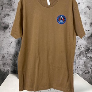 May include: A tan crew neck t-shirt with a small circular emblem on the upper left chest. The emblem features a bird, the text "PHOENIX RAVEN", and "GLOBAL PROTECTION" in blue and red. The shirt is hanging on a black hanger.