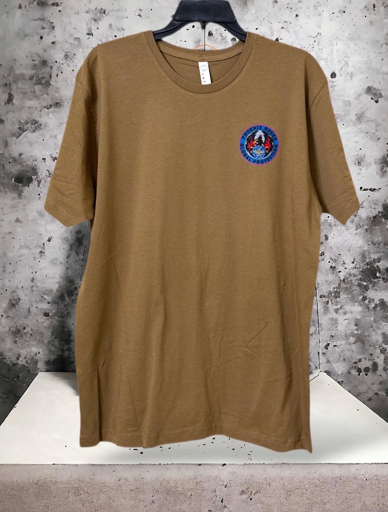 May include: A tan crew neck t-shirt with a small circular emblem on the upper left chest. The emblem features a bird, the text "PHOENIX RAVEN", and "GLOBAL PROTECTION" in blue and red. The shirt is hanging on a black hanger.