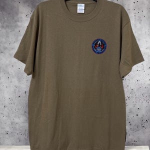 May include: Olive green t-shirt with a crew neck and short sleeves. A circular emblem with the text "Phoenix Raven Global Protection" is on the left chest. The shirt is hanging on a black hanger against a speckled gray background.