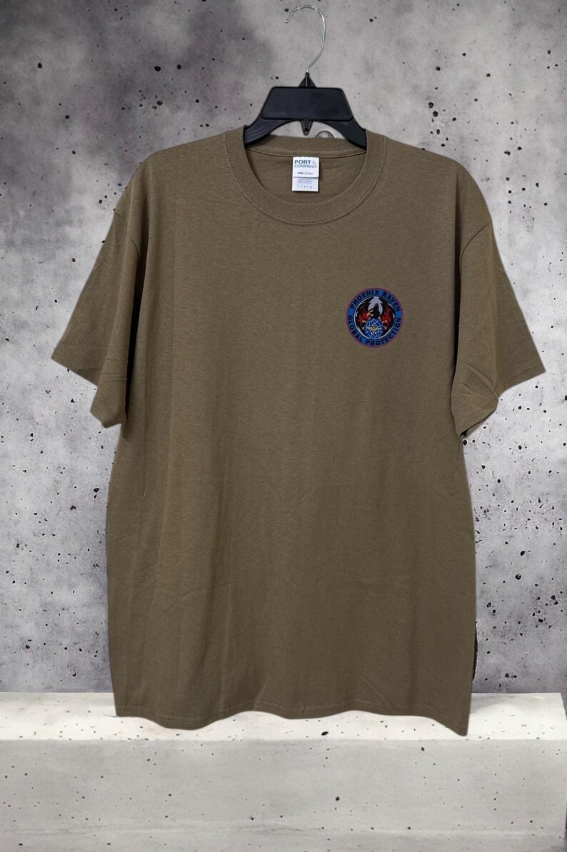 May include: Olive green t-shirt with a crew neck and short sleeves. A circular emblem with the text "Phoenix Raven Global Protection" is on the left chest. The shirt is hanging on a black hanger against a speckled gray background.