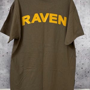 May include: Olive green t-shirt with the word "RAVEN" printed in yellow across the back. The shirt is hanging on a hanger against a mottled gray and white background. The short-sleeved shirt is made of a soft material.