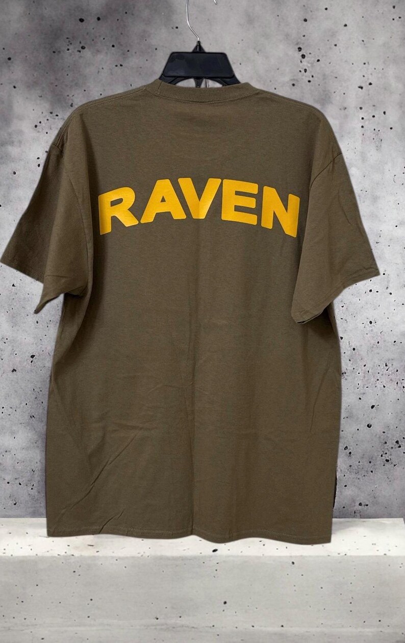 May include: Olive green t-shirt with the word "RAVEN" printed in yellow across the back. The shirt is hanging on a hanger against a mottled gray and white background. The short-sleeved shirt is made of a soft material.