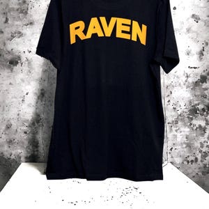 May include: Black t-shirt with the word "RAVEN" printed in yellow block letters across the back. The shirt is hanging on a black hanger against a mottled gray and white background. The shirt is displayed on a white surface.