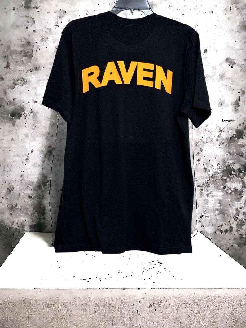 May include: Black t-shirt with the word "RAVEN" printed in yellow block letters across the back. The shirt is hanging on a black hanger against a mottled gray and white background. The shirt is displayed on a white surface.