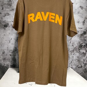 May include: A brown t-shirt with the word "RAVEN" printed in bright orange across the back. The shirt is made of a soft material and has a classic crew neck design. The shirt is hanging on a black hanger.