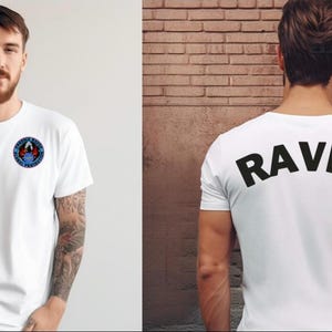 May include: White t-shirts with graphic designs. One shirt has a circular emblem on the front. The other shirt has the word "RAVEN" printed in large black letters across the back. Both shirts are short-sleeved.