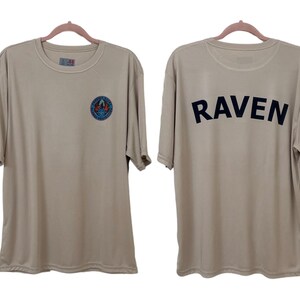 May include: Two-sided tan t-shirt. The front features a circular emblem with blue and orange accents. The back displays the word "RAVEN" in bold, black capital letters. The shirt is hanging on a pink hanger.