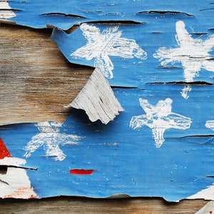 May include: Close-up of a weathered wooden surface with peeling blue paint. The paint is chipped and cracked, revealing the wood underneath. White stars are painted on the blue surface, representing the American flag.