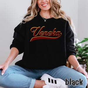 A person wearing a black crewneck sweatshirt with "Veneka" in red and yellow script across the chest, with "black" written in white in the bottom right corner.