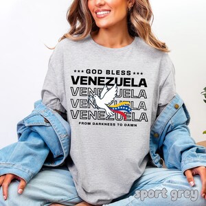 May include: A light gray long-sleeve shirt with the text "GOD BLESS VENEZUELA" in black. A white dove with the Venezuelan flag is featured. The shirt also has the phrase "FROM DARKNESS TO DAWN".