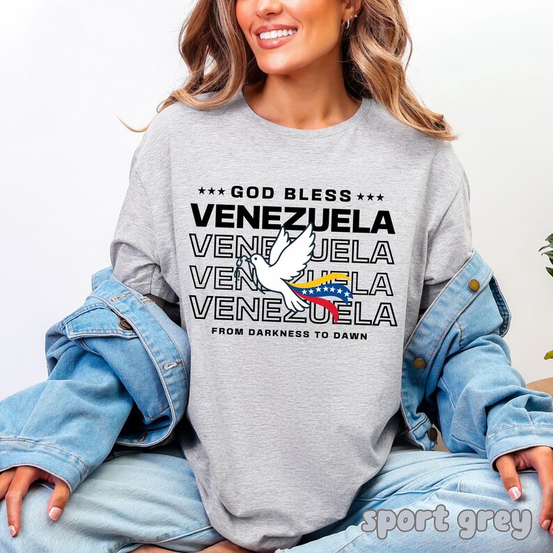 May include: A light gray long-sleeve shirt with the text "GOD BLESS VENEZUELA" in black. A white dove with the Venezuelan flag is featured. The shirt also has the phrase "FROM DARKNESS TO DAWN".