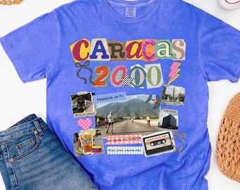 Può includere: Una maglietta blu con un design grafico con la parola "CARACAS" in lettere colorate, il numero "2000" e varie immagini. La maglietta ha anche il testo "FLO BLUE" in basso.