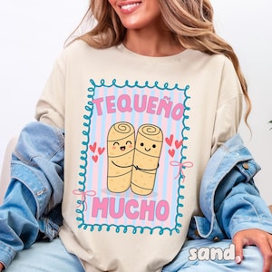 Könnte beinhalten: Sandfarbenes Sweatshirt mit einer Grafik, die zwei lächelnde Cartoon-Tequeños zeigt, darüber der Text "TEQUEÑO" und darunter "MUCHO". Das Design wird von einem blauen Rahmen mit rosa Herzen und Akzenten umrandet.