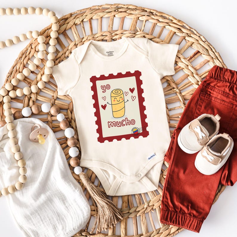 May include: Cream-colored baby onesie with a red postage stamp design featuring a cartoon arepa and the words "yo mucho." The onesie is paired with rust-colored pants and white and tan baby shoes. A pacifier and wooden beads are also in the image.