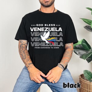 May include: Black t-shirt with the text "GOD BLESS VENEZUELA" and a white dove graphic holding the Venezuelan flag. The phrase "FROM DARKNESS TO DAWN" is printed below the text. The word "black" is printed at the bottom right.