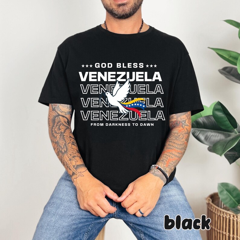 May include: Black t-shirt with the text "GOD BLESS VENEZUELA" and a white dove graphic holding the Venezuelan flag. The phrase "FROM DARKNESS TO DAWN" is printed below the text. The word "black" is printed at the bottom right.