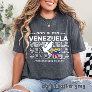 May include: Dark heather grey t-shirt with the text "GOD BLESS VENEZUELA" and a dove graphic holding a Venezuelan flag. The shirt also includes the phrase "FROM DARKNESS TO DAWN".