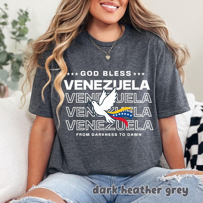 May include: Dark heather grey t-shirt with the text "GOD BLESS VENEZUELA" and a dove graphic holding a Venezuelan flag. The shirt also includes the phrase "FROM DARKNESS TO DAWN".