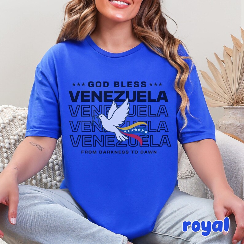 May include: Royal blue t-shirt with the text "GOD BLESS VENEZUELA" in black. A white dove with a Venezuelan flag is in the center. The phrase "FROM DARKNESS TO DAWN" is below the text.