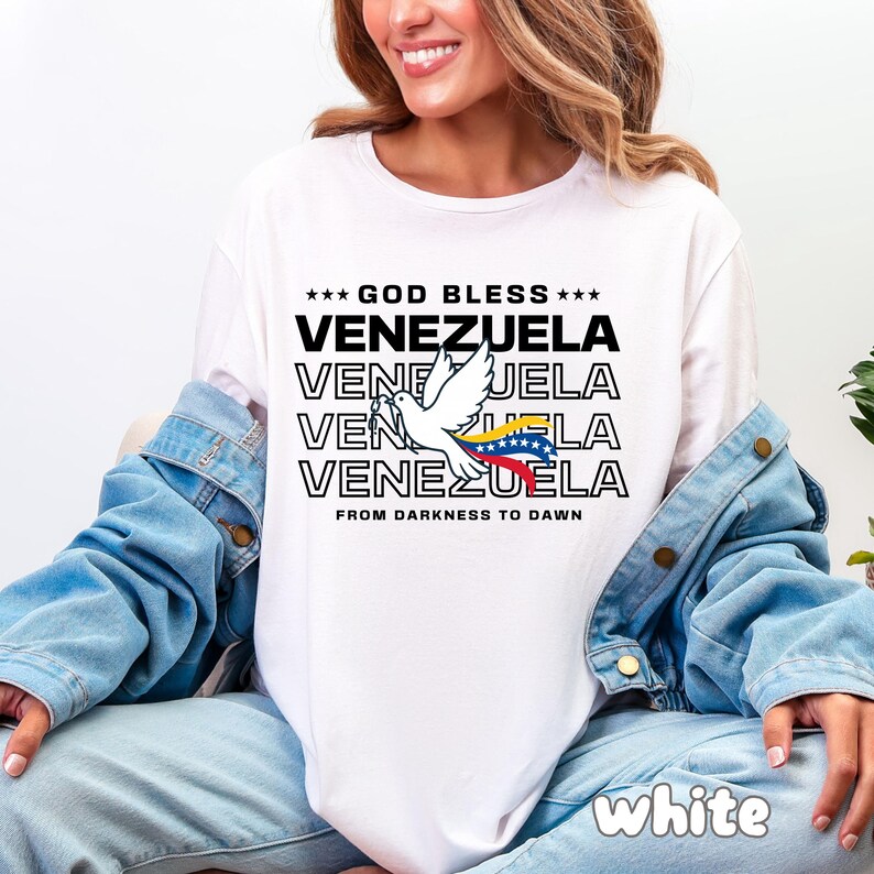 May include: White t-shirt with the text "GOD BLESS VENEZUELA" in black, stacked vertically. A white dove with a Venezuelan flag is in the center. The phrase "FROM DARKNESS TO DAWN" is below the text. The word "white" is at the bottom.
