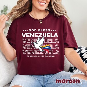 May include: Maroon t-shirt with the text "GOD BLESS VENEZUELA" repeated four times, featuring a white dove and the Venezuelan flag. The phrase "FROM DARKNESS TO DAWN" is printed below the text. The word "maroon" is printed at the bottom right.