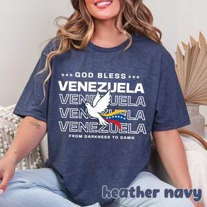 May include: Heather navy t-shirt with the text "GOD BLESS VENEZUELA" in white, stacked vertically. A white dove with a Venezuelan flag design is featured. The phrase "FROM DARKNESS TO DAWN" is below the text. The words "heather navy" are at the bottom.