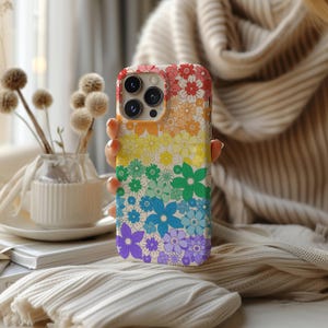 Puede incluir: Una funda blanca para iPhone con un patrón floral de arcoíris. La funda tiene un recorte para el objetivo de la cámara y un fondo blanco.