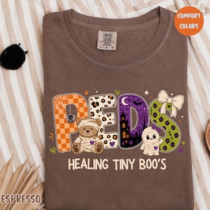 May include: A brown crewneck t-shirt featuring the text PEDS HEALING TINY BOO'S. The letters are decorated with orange checkerboard, leopard print, purple haunted house, and green patterns. A mummy teddy bear and a mummy ghost are included.