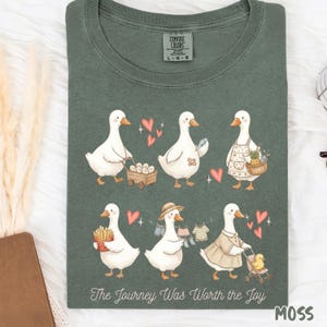 Könnte beinhalten: Moosgrünes T-Shirt mit einer skurrilen Illustration weißer Gänse. Die Gänse sind mit Herzen dargestellt und tragen Gegenstände wie Pommes Frites, einen Obstkorb und einen Kinderwagen. Der Text "The Journey Was Worth the Joy" ist unten aufgedruckt.