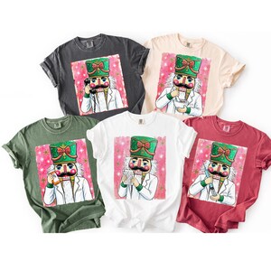 May include: Five short-sleeved t-shirts in charcoal, cream, olive green, white, and maroon. Each shirt features a graphic of a nutcracker dressed as a medical professional, holding items like a telephone, mortar and pestle, pill bottle, or syringe.