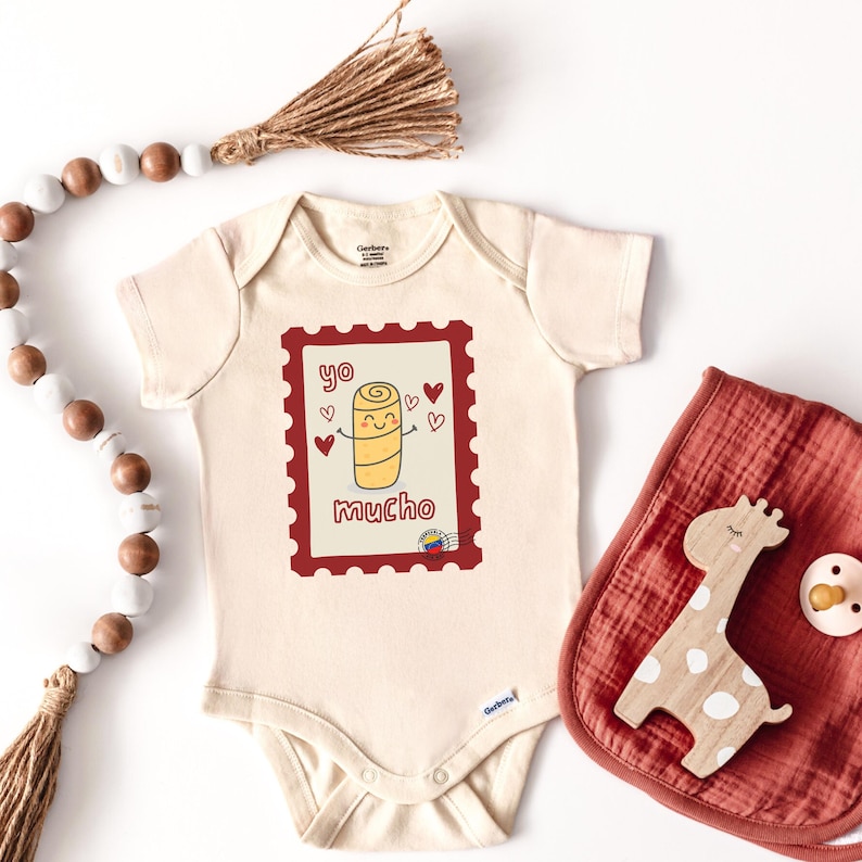 May include: Cream-colored baby onesie with a red postage stamp design featuring a cartoon arepa and the words "yo mucho." A red bib with a wooden giraffe toy and a pacifier are next to the onesie.