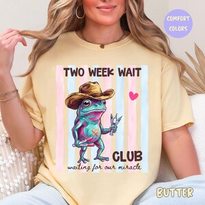 以下が含まれることがあります： バター色のTシャツ。カウボーイハットをかぶったカエルのグラフィック、注射器と包帯を持っています。「TWO WEEK WAIT CLUB waiting for our miracle」の文字とピンクと青のストライプ。